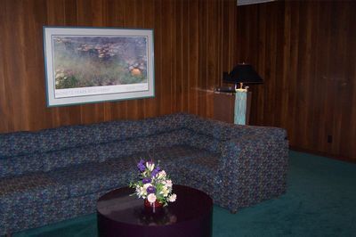 Blue patterned sectional sofa in a wood-paneled room with a framed painting, lamp, and small flower arrangement on a table