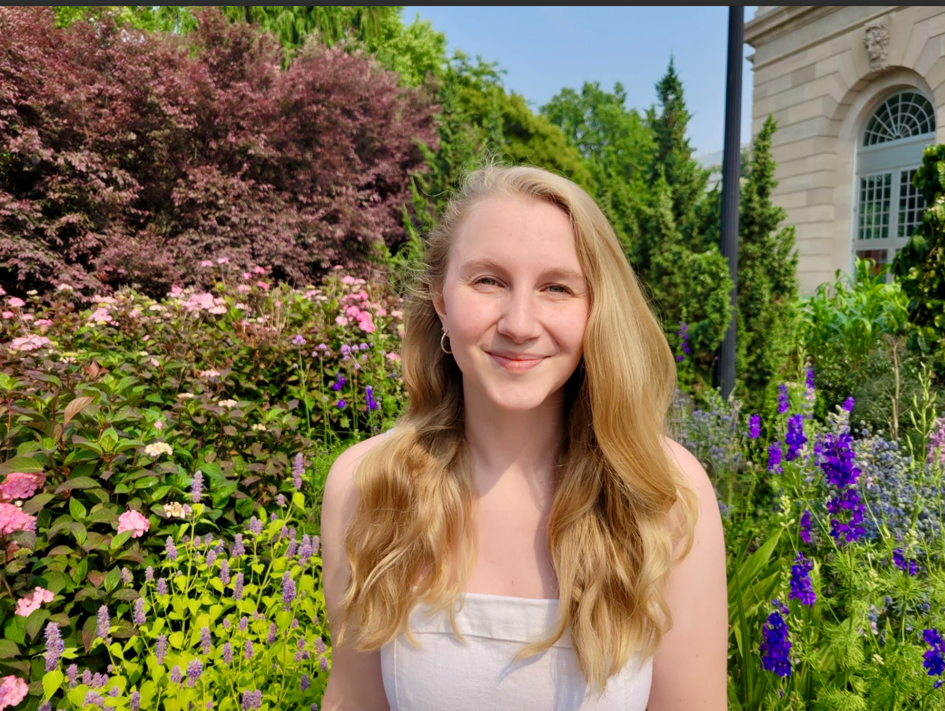 Woman with blonde hair smiling in a garden with colorful flowers and trees.