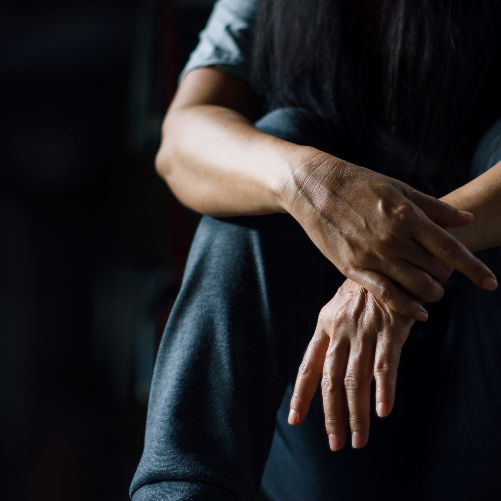 Woman sits with head down, hands clasped. Dark setting suggests isolation or sadness.