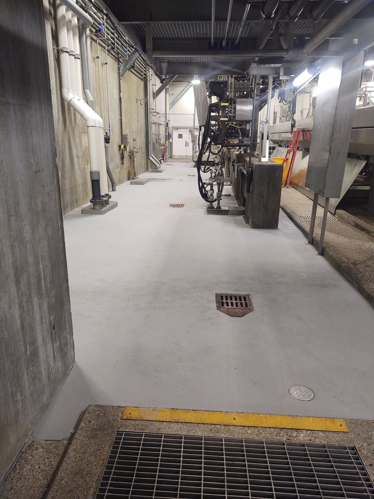 Industrial corridor with concrete floor, overhead pipes, and metal grates in a dimly lit facility