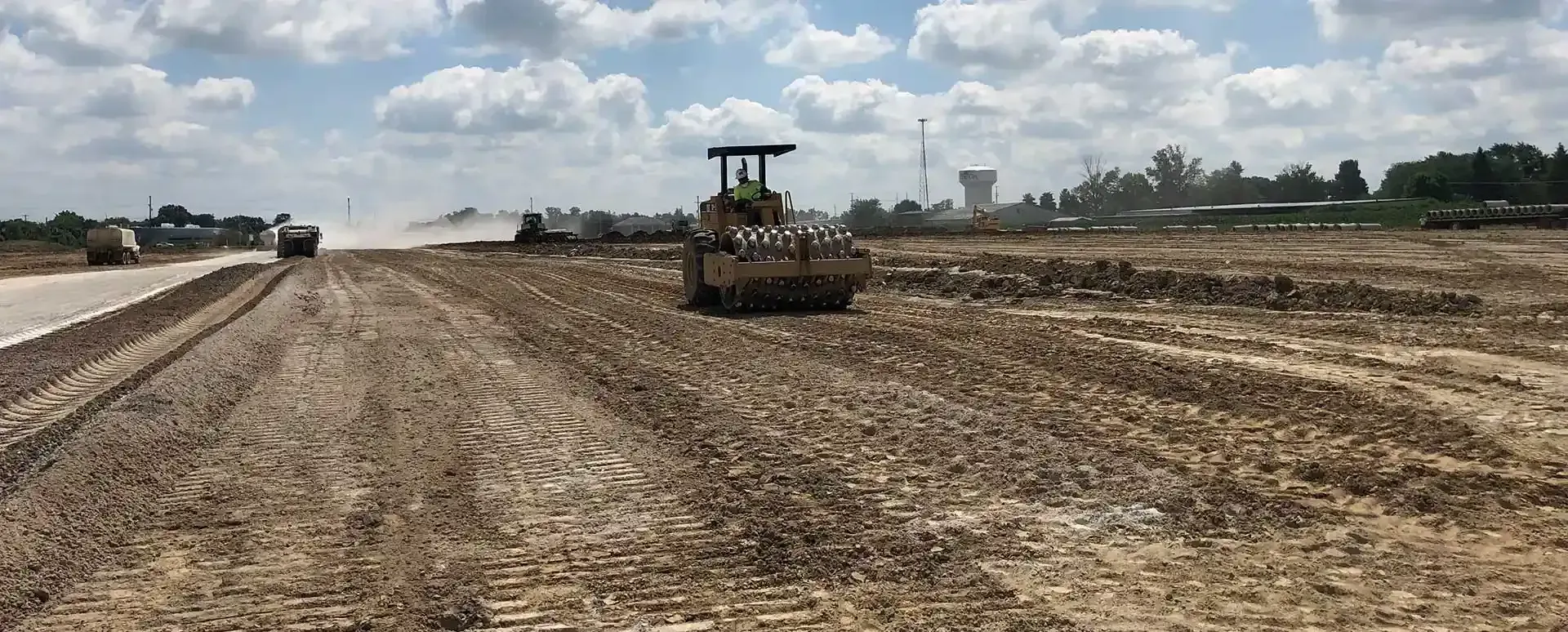 Bulldozer grading a dirt field beside a road under a partly cloudy sky