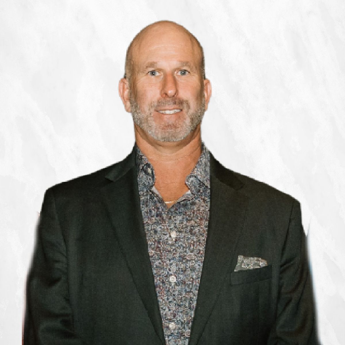Man in a dark blazer and patterned shirt smiling against a white background