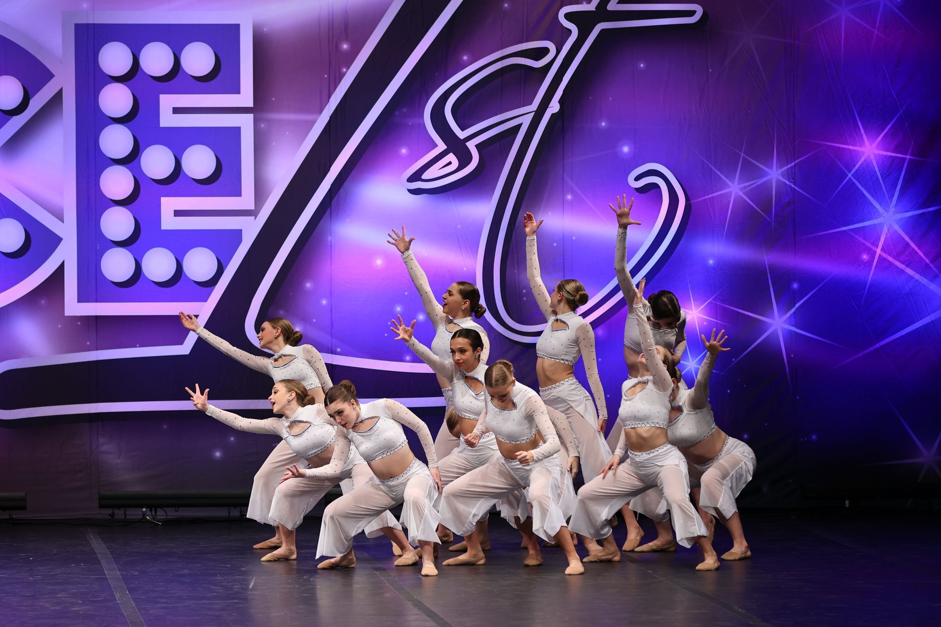 Dance troupe in white costumes striking a synchronized pose on a purple-lit stage