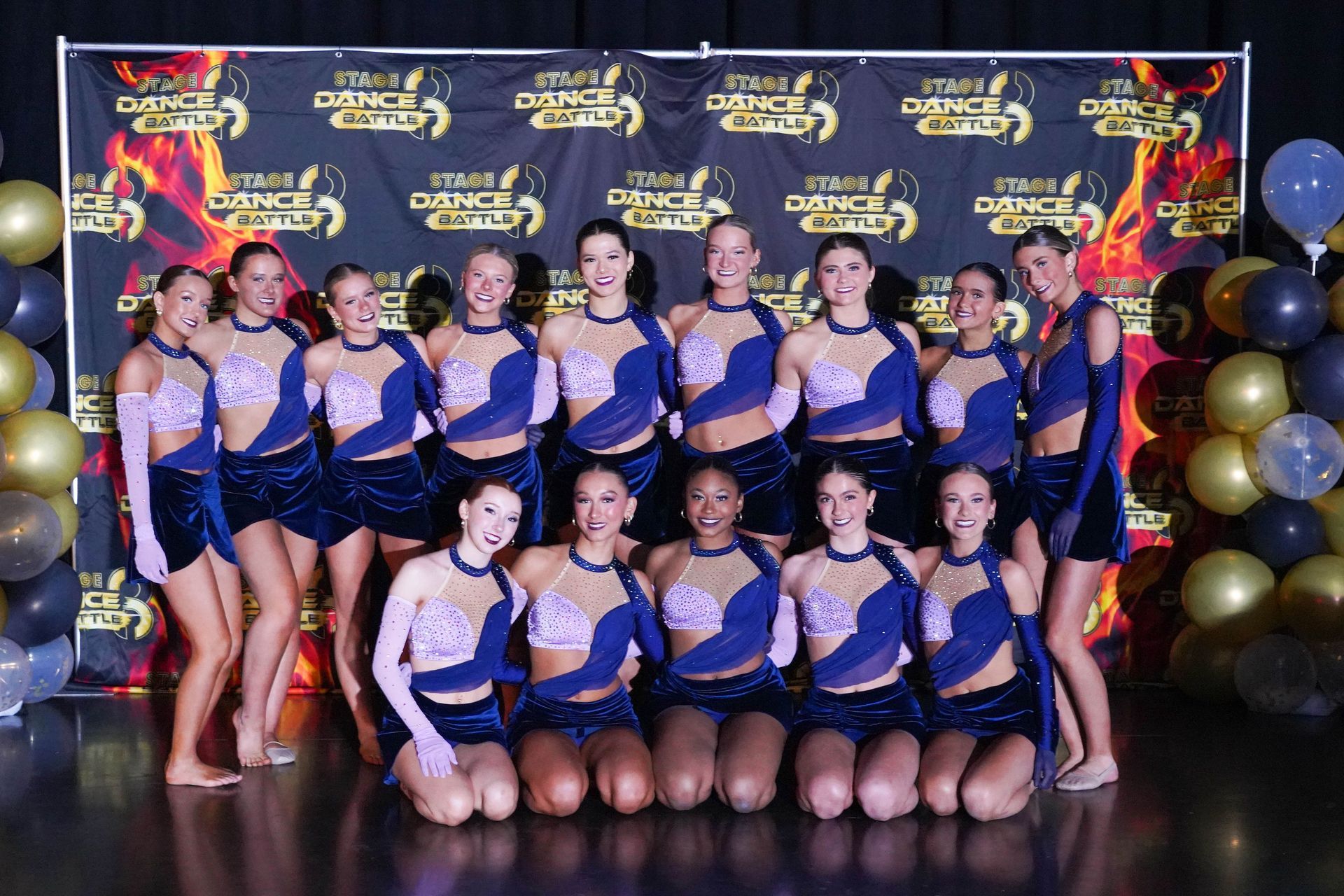 Cheer squad posing in blue-and-gold uniforms at a competition backdrop with balloons