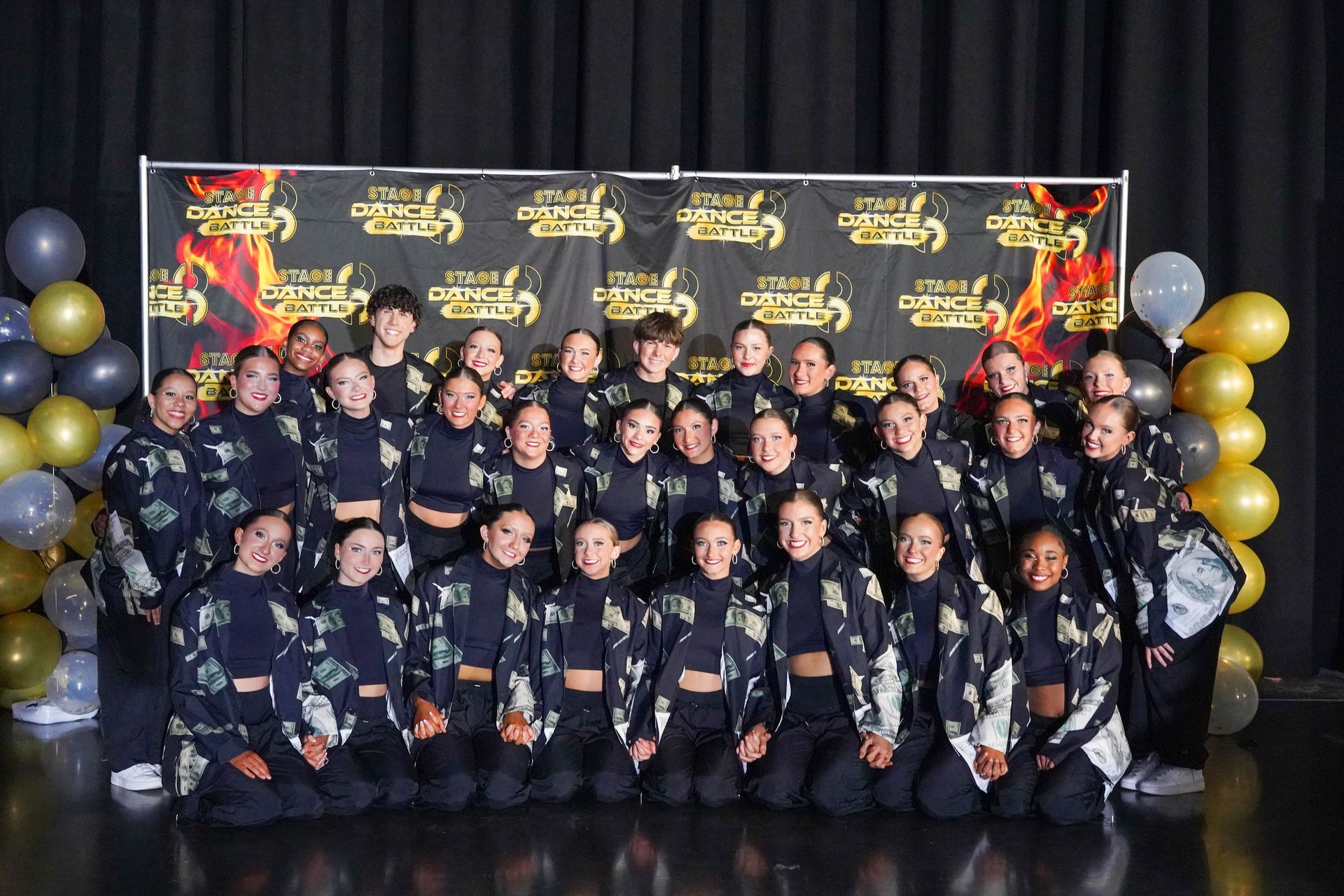 Group of dancers posing in matching black outfits in front of a gold-and-black event backdrop with balloons