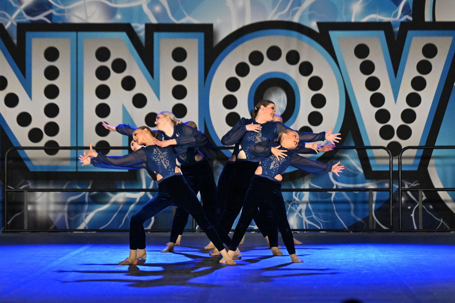 Dancers in blue costumes performing on a stage with a large "NOW" backdrop