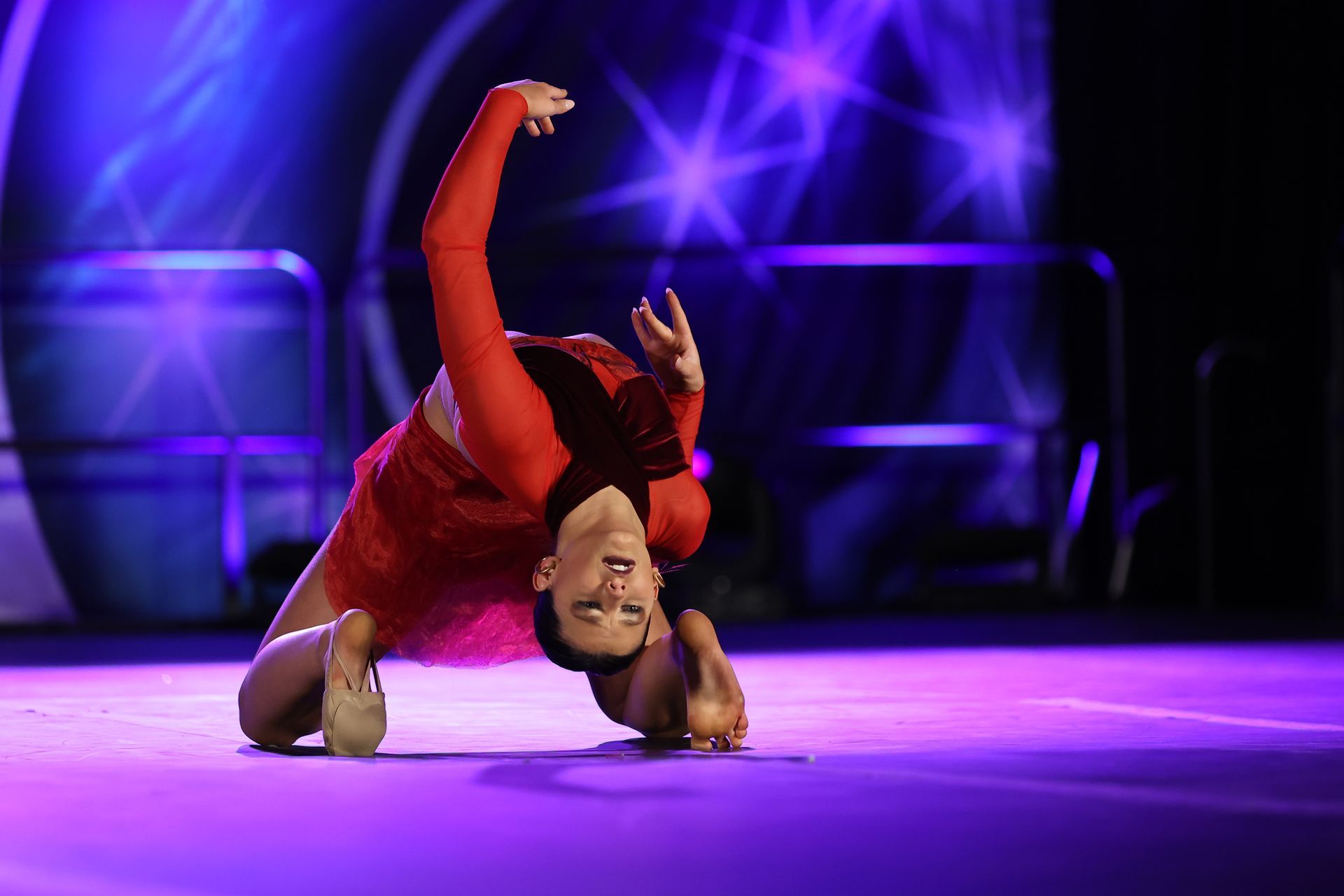 Dancer in red costume performing an acrobatic floor pose on a purple-lit stage