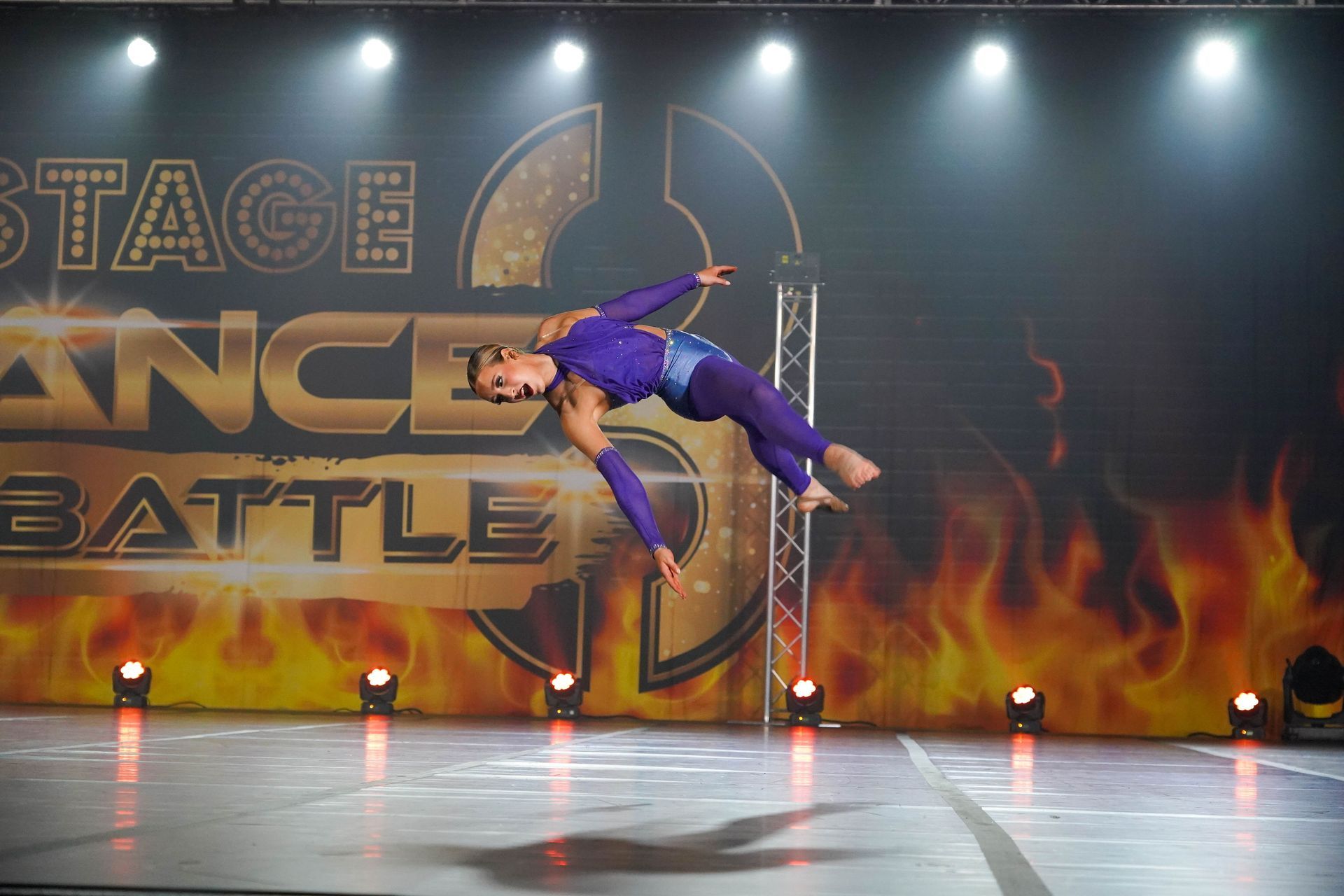 Contemporary dancer in purple midair on stage with flames and “Stage Dance Battle” backdrop