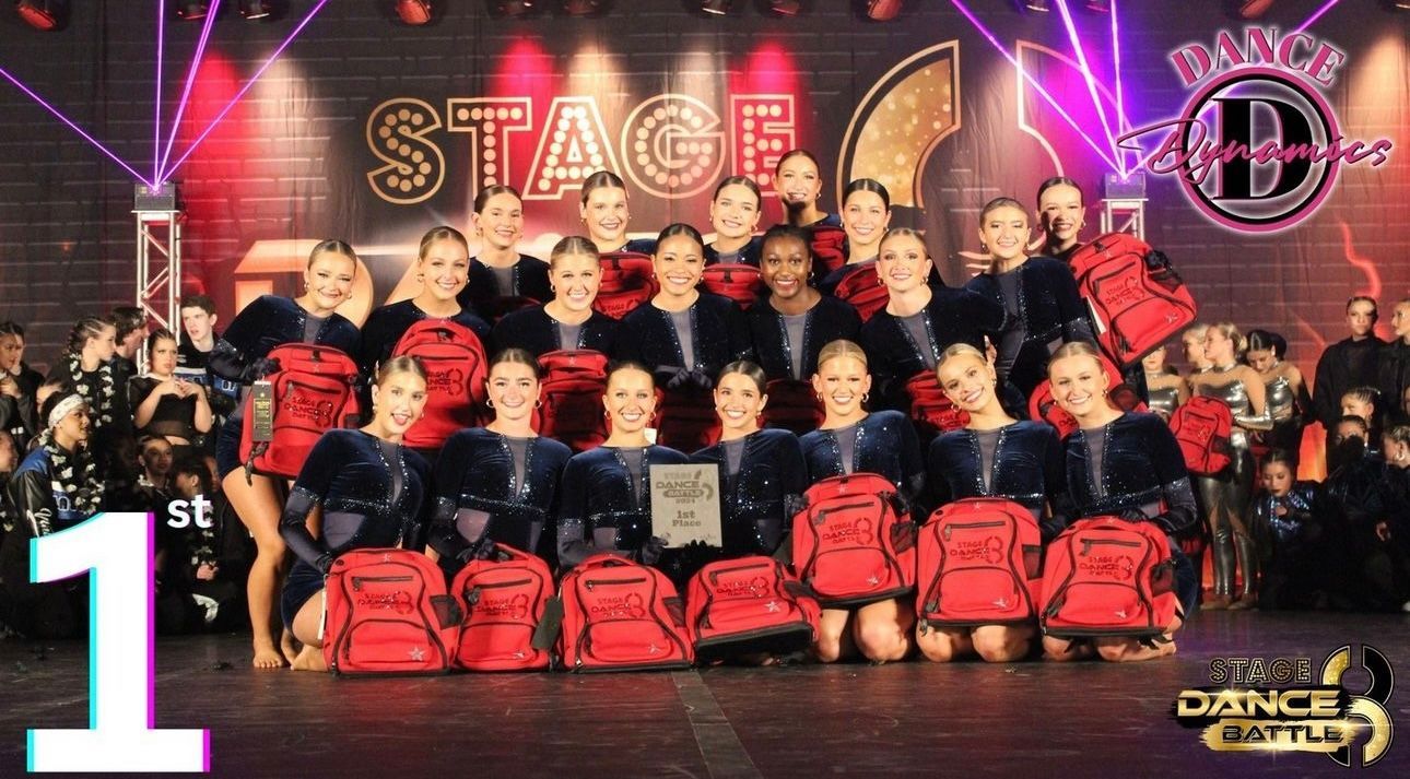 Group dance team posing on stage with red duffel bags and pink neon lights, “STAGE” sign behind them.