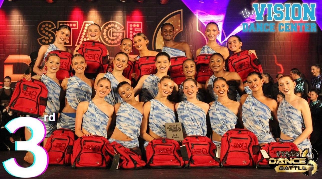 Group of performers posing on stage in red costumes, holding red props with colorful stage lights behind them.
