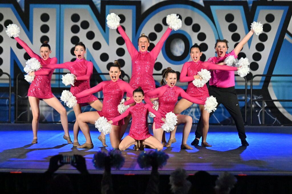 Group of dancers in pink costumes performing on a stage with large “WOW” letters behind them
