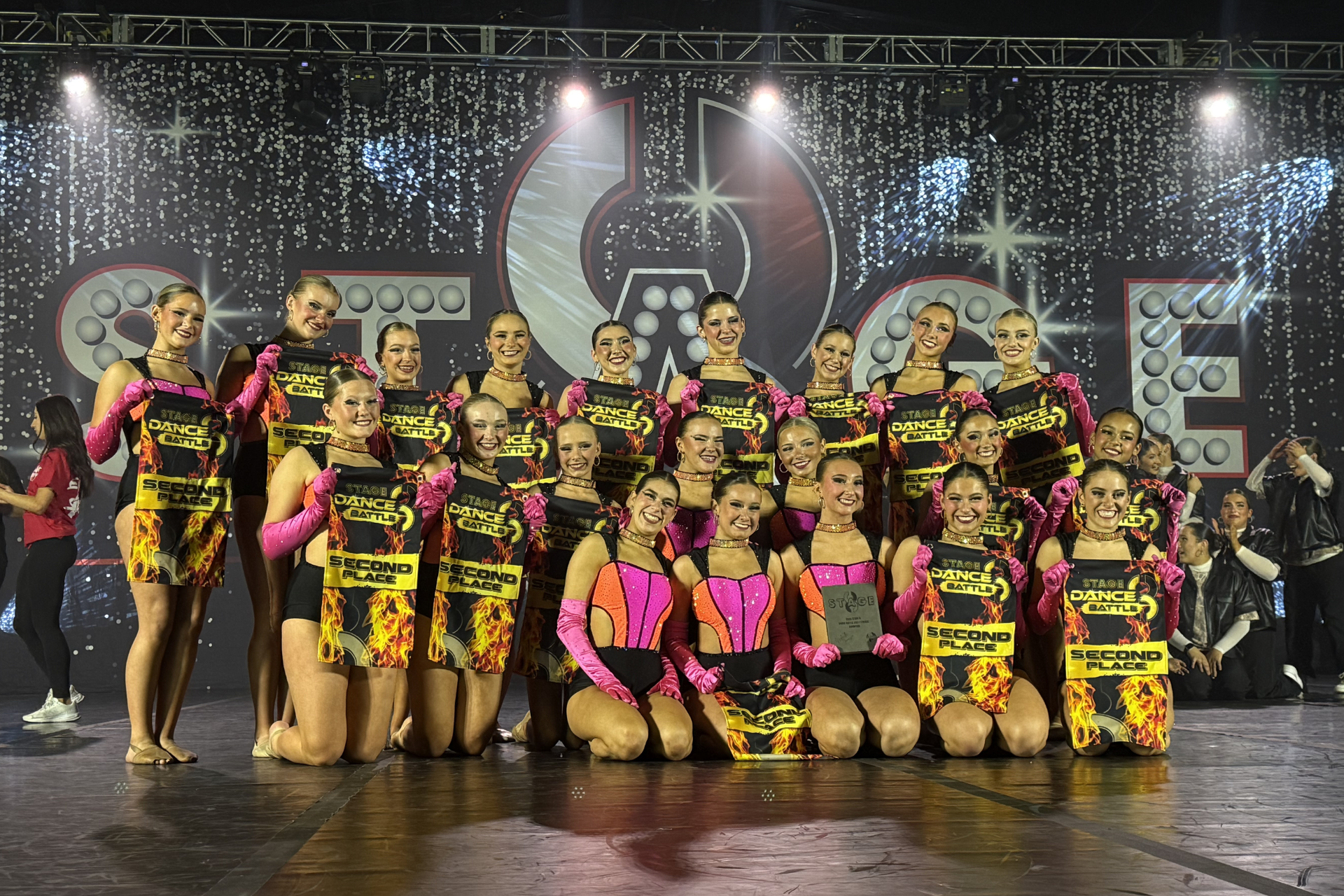 Group of dancers in black and pink costumes posing on a stage with bright lights and a large backdrop