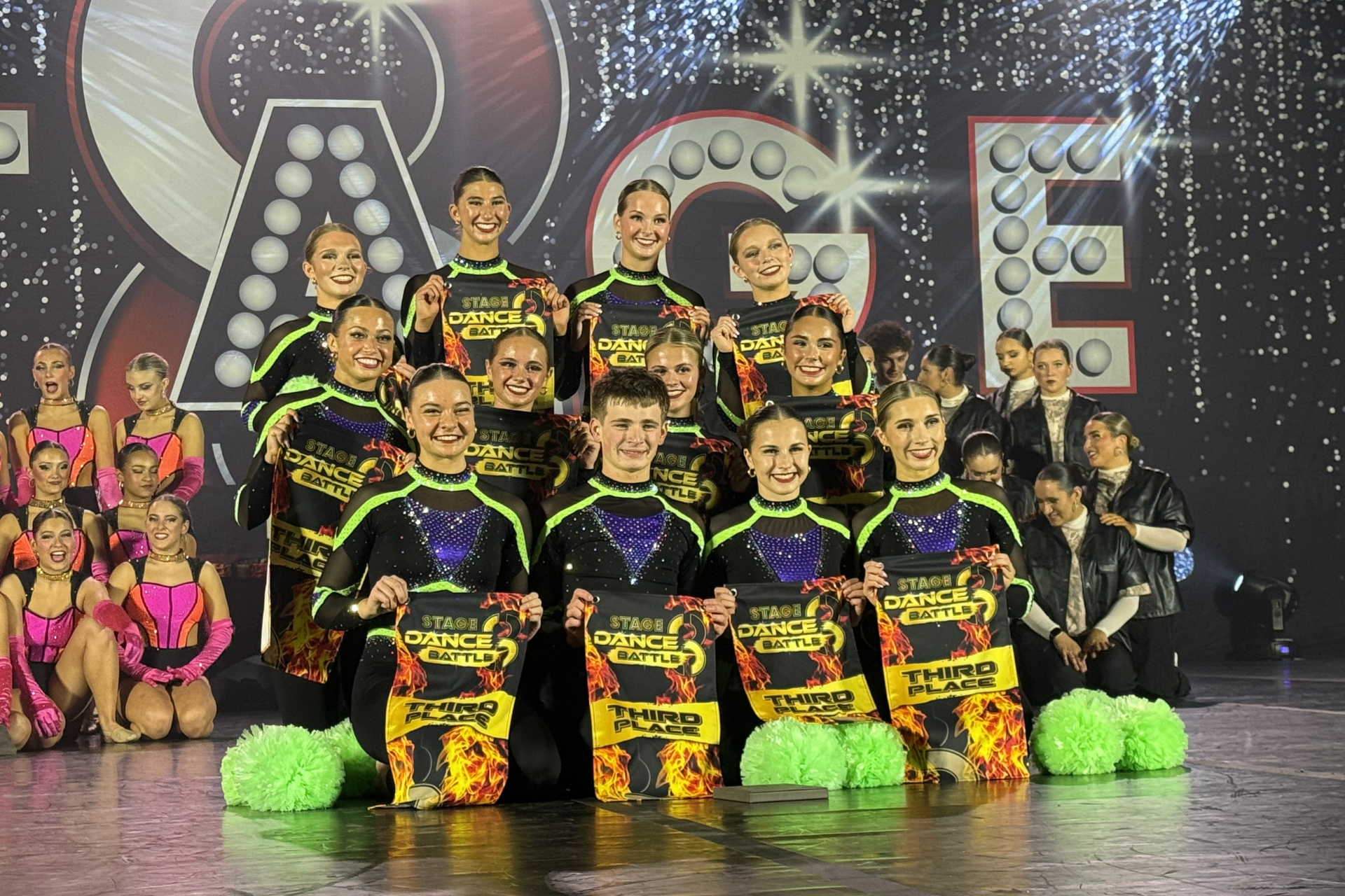 Cheer team posing on stage in black, neon green, and yellow uniforms with sparkly backdrop and pom-poms