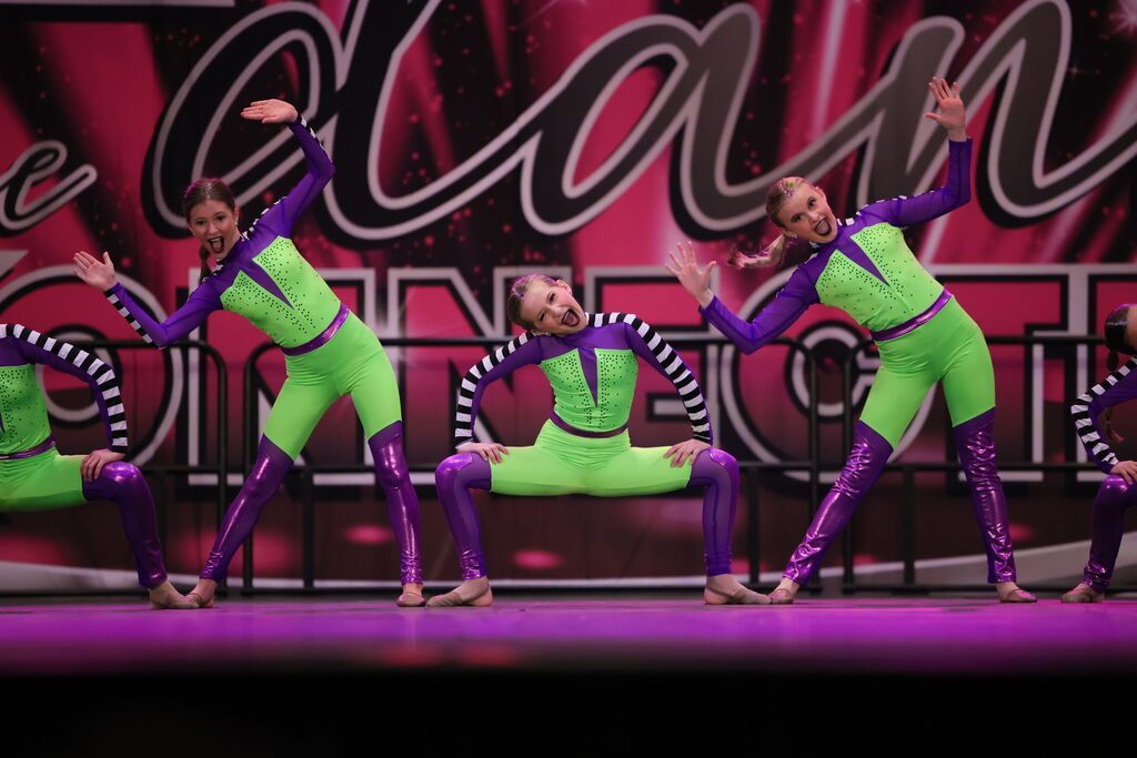 Three dancers in neon green and purple costumes pose on a stage under pink lighting.
