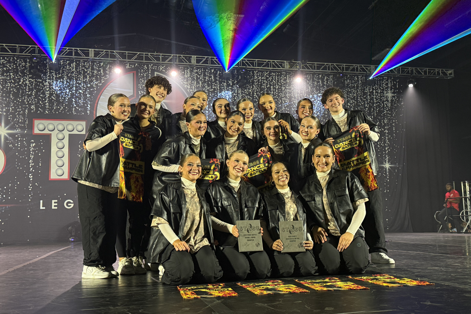 Group of dancers posing onstage in matching black and gold outfits under colorful laser lights