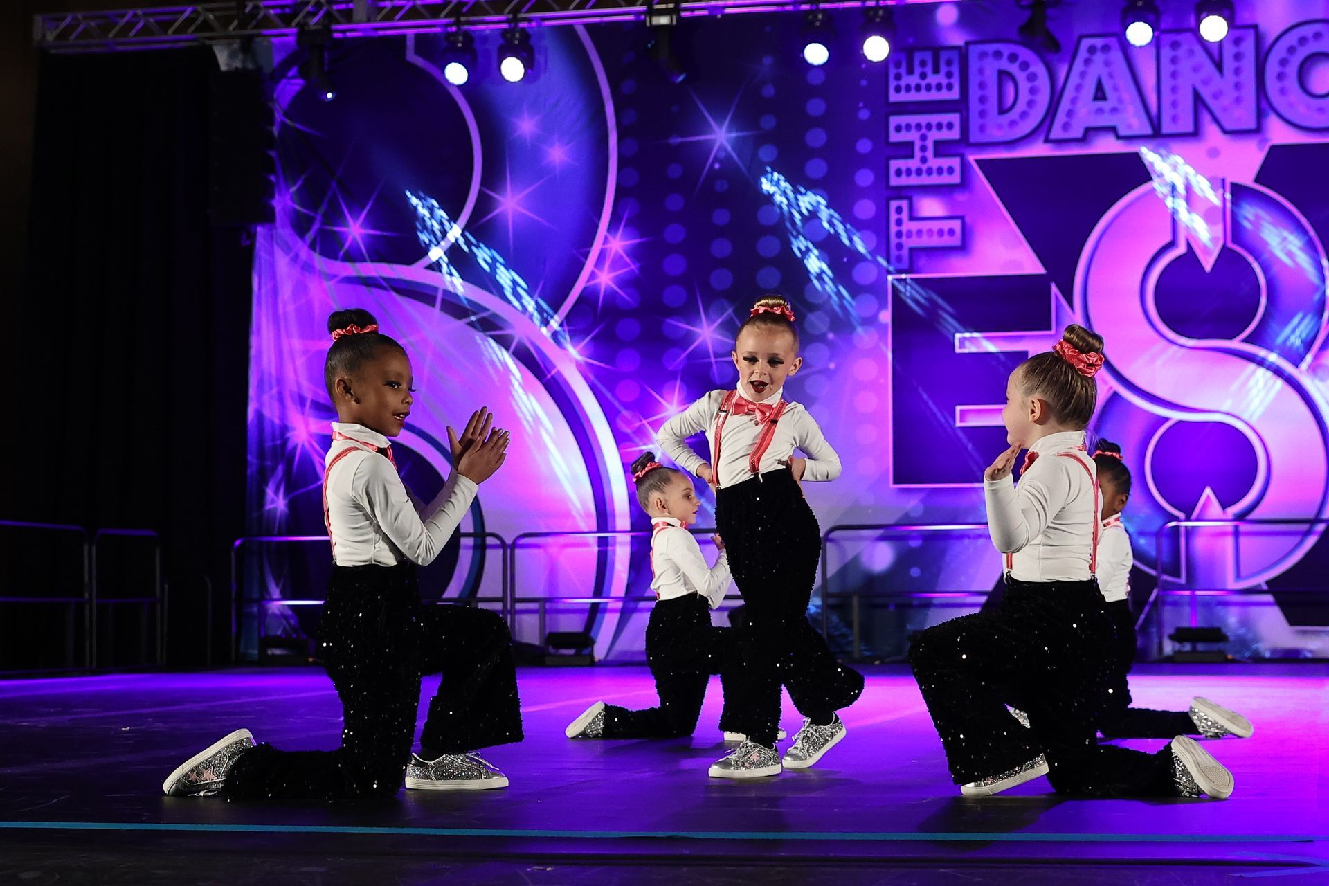 Three dancers kneel on stage under purple lights with a neon dance backdrop.