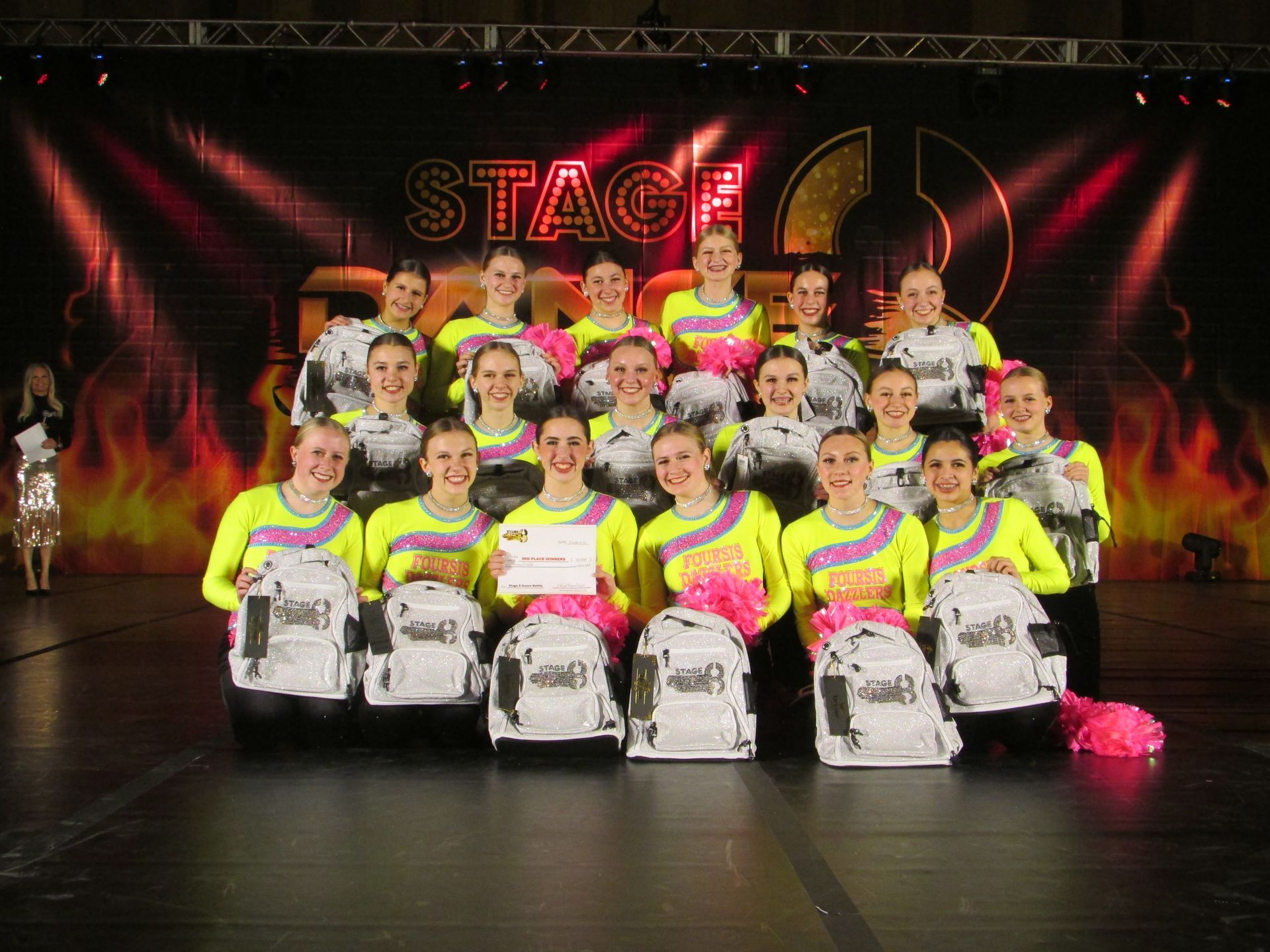 Group of performers in neon yellow costumes posing on a lit stage with drums and a red “STAGE” backdrop