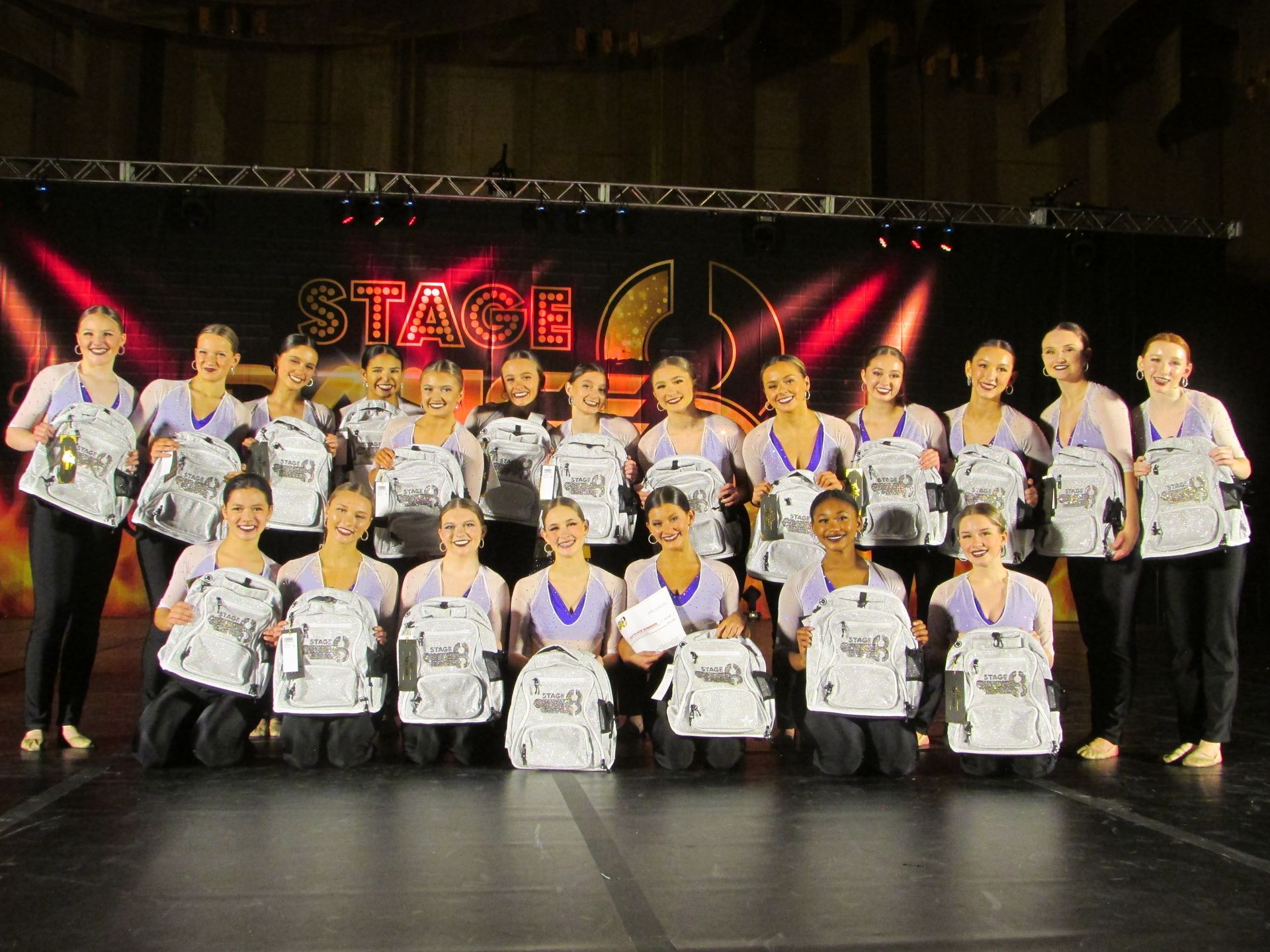 Dance team posing on stage with trophies, wearing matching white shirts and black pants under red lights