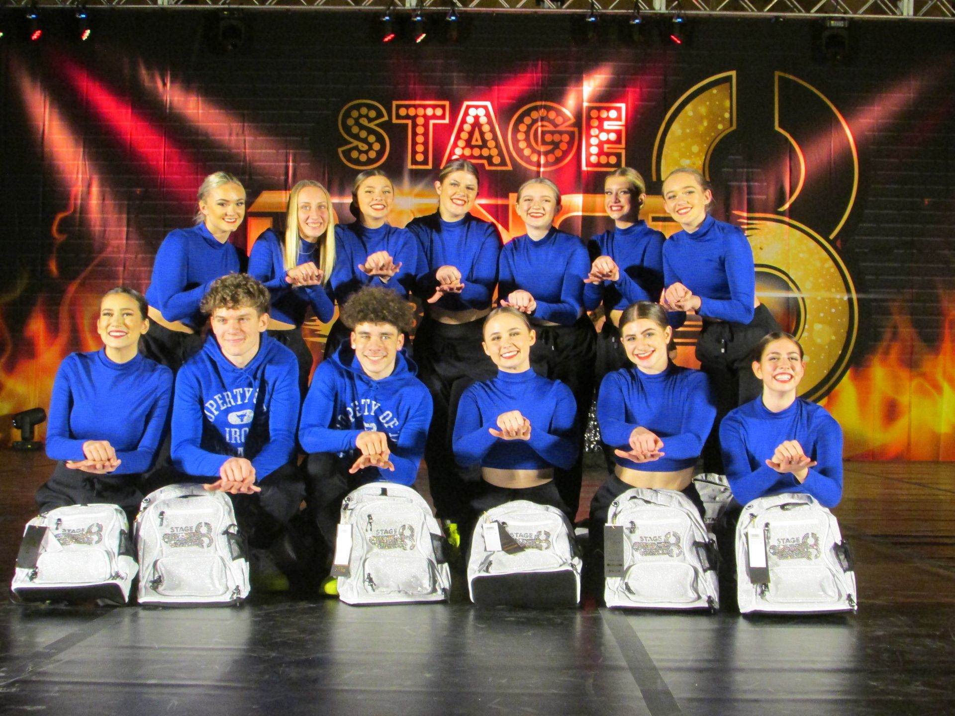 Group of performers in blue outfits posing on stage in front of a glowing “STAGE” backdrop