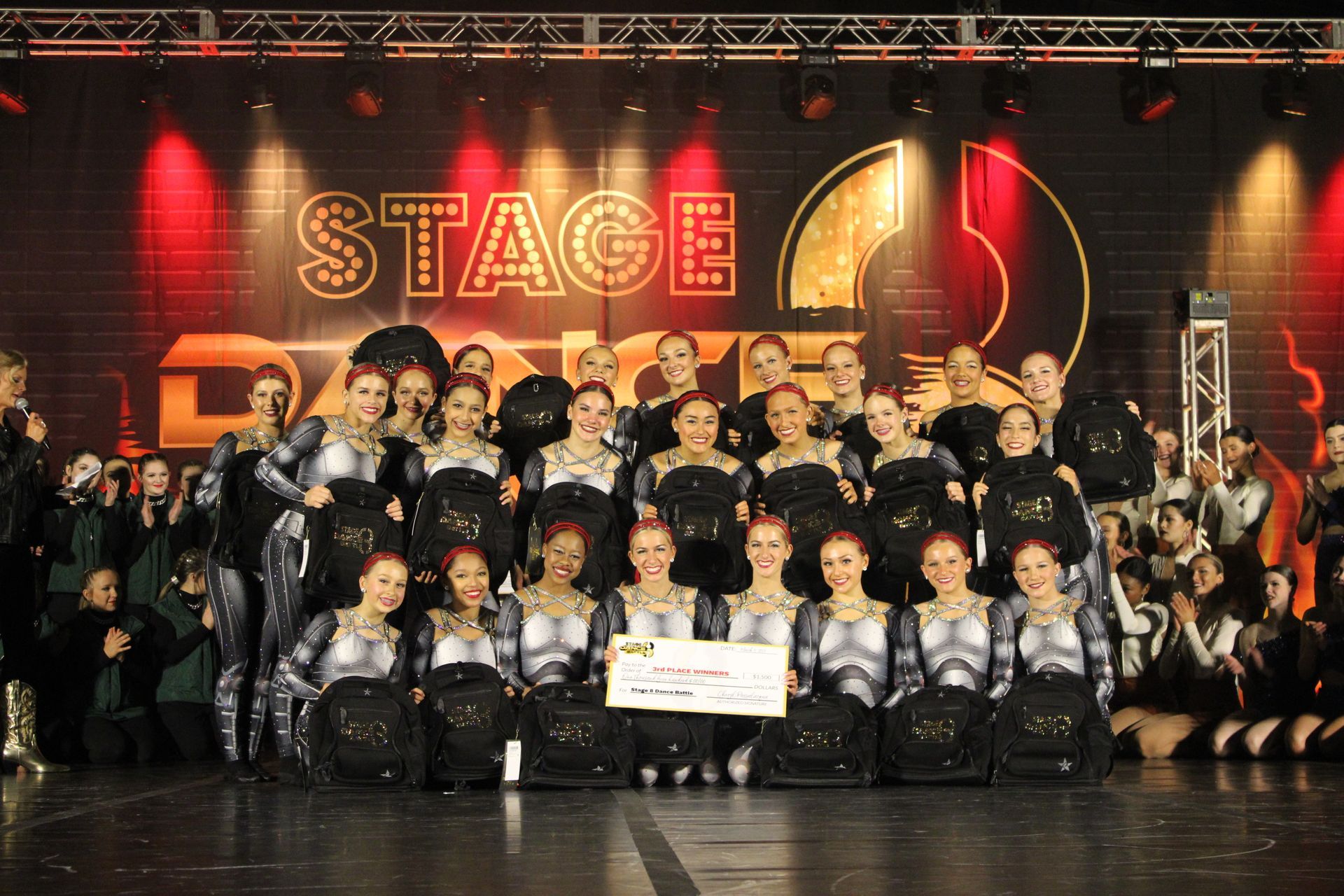 Large dance troupe posing on stage in black costumes with a STAGE sign and spotlight backdrop