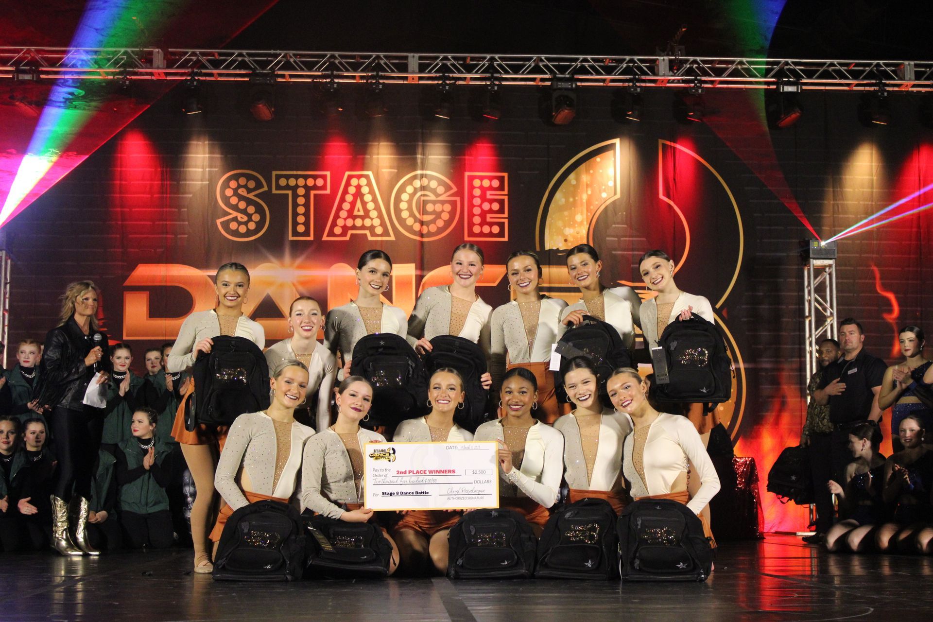 Group of performers posing on a lit stage with a “STAGE” backdrop, holding a sign, in front of bright red lights