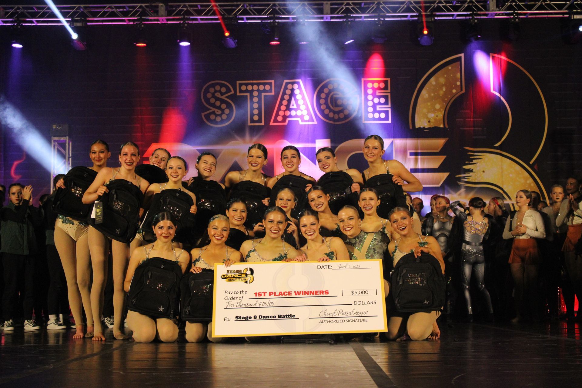 Dance troupe posing onstage with a large check in front of a “STAGE” backdrop