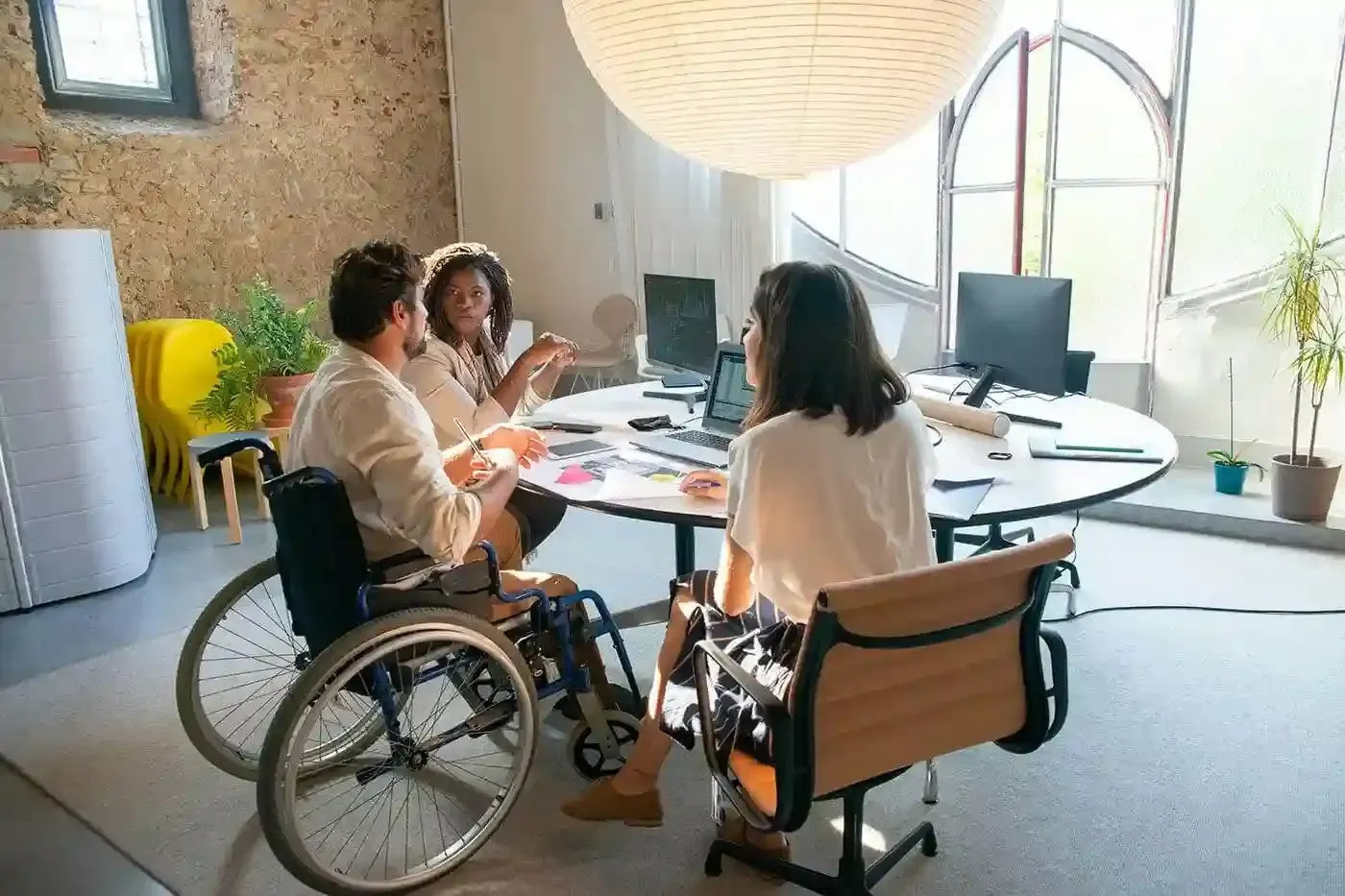 People in wheelchairs and a seated colleague in a bright office meeting around a desk
