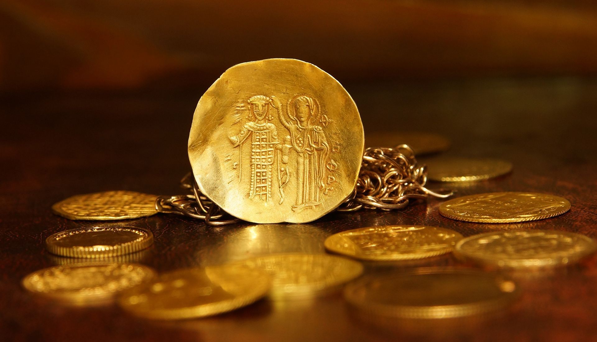Gold coins on a reflective surface, one coin upright showing a carved design, surrounded by a gold chain and other coins.