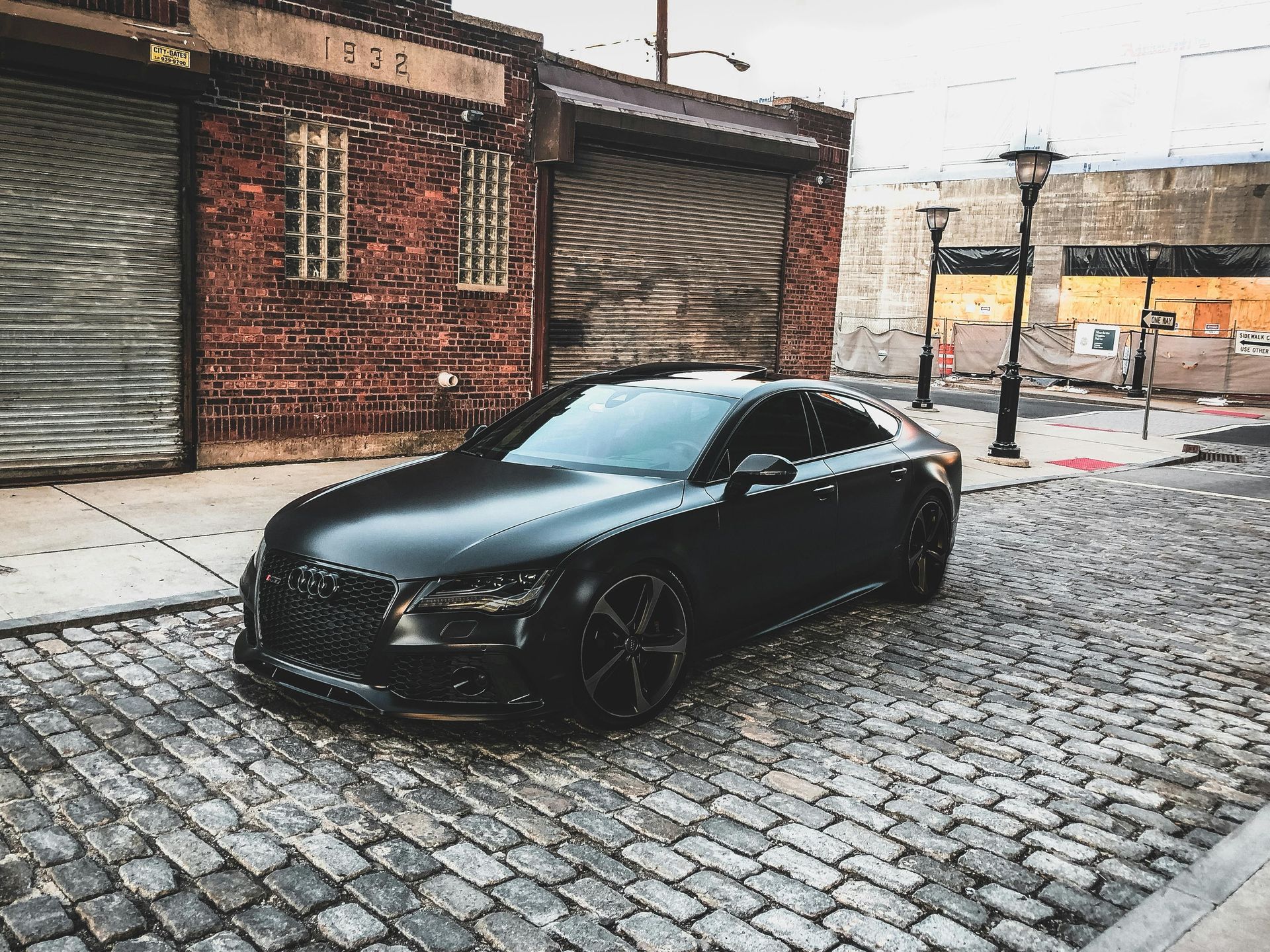 A black car is parked on a cobblestone street in front of a brick building. - abraham sanieoff