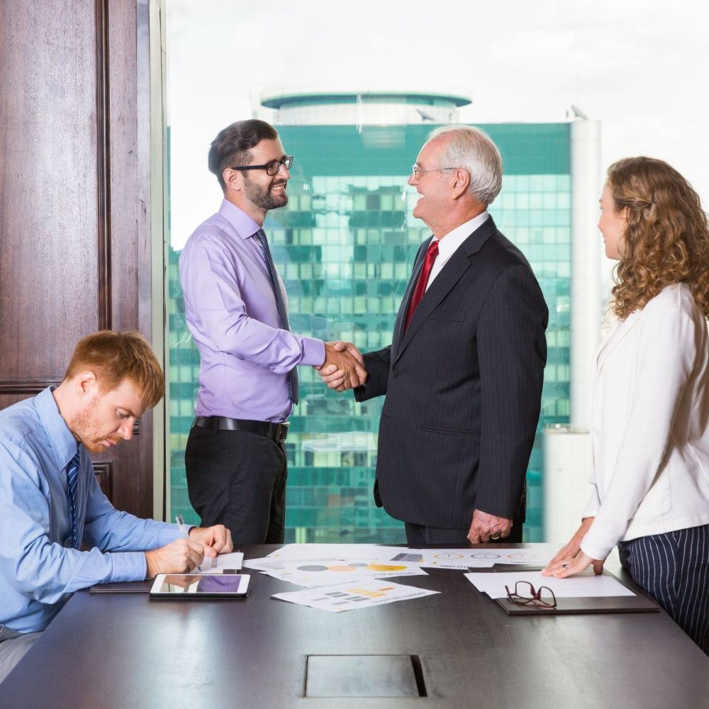 Businesspeople shaking hands in an office, others watching and working at a table.