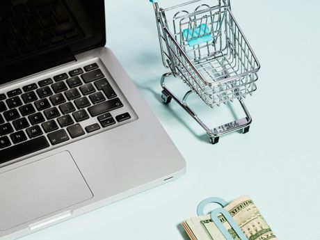 A shopping cart is sitting on top of a laptop next to a stack of money.