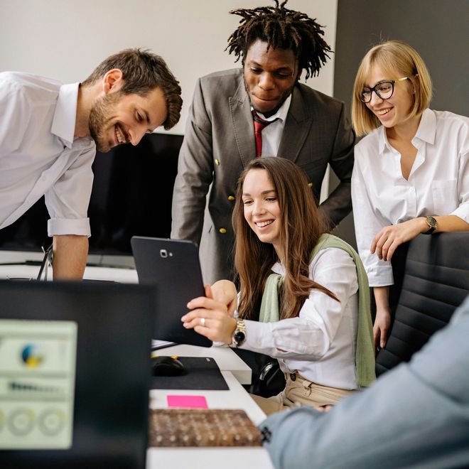 Team of five colleagues gathered around a laptop at an office desk, smiling and collaborating.
