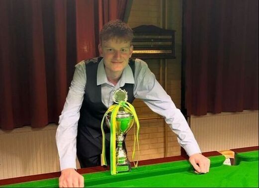 Jongeman in vest lacht, houdt een trofee vast op een snookertafel, groen laken, houten paneelachtergrond.