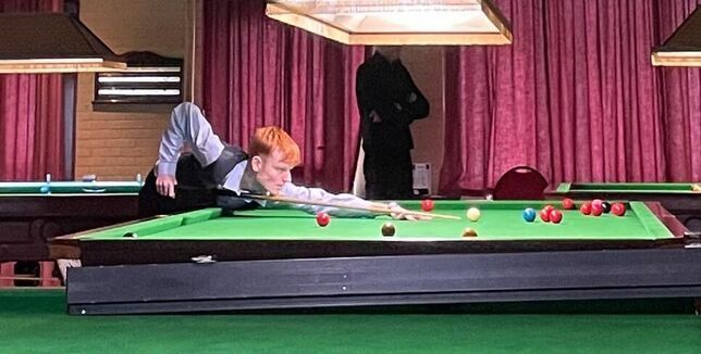 Een jongeman met rood haar speelt snooker op een groene tafel in een club, met rode gordijnen op de achtergrond.