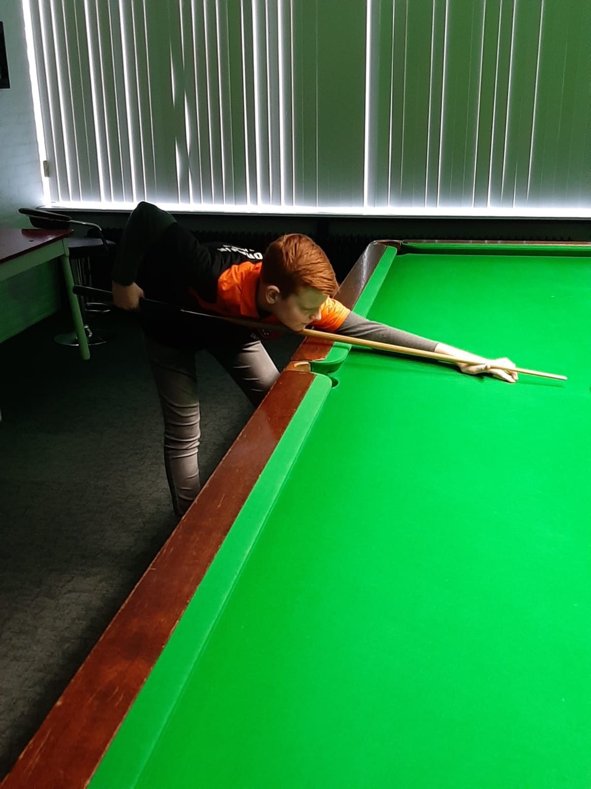 Een jongeman met kastanjebruin haar bereidt een bal voor op een groene snookertafel, met een keu in de hand.