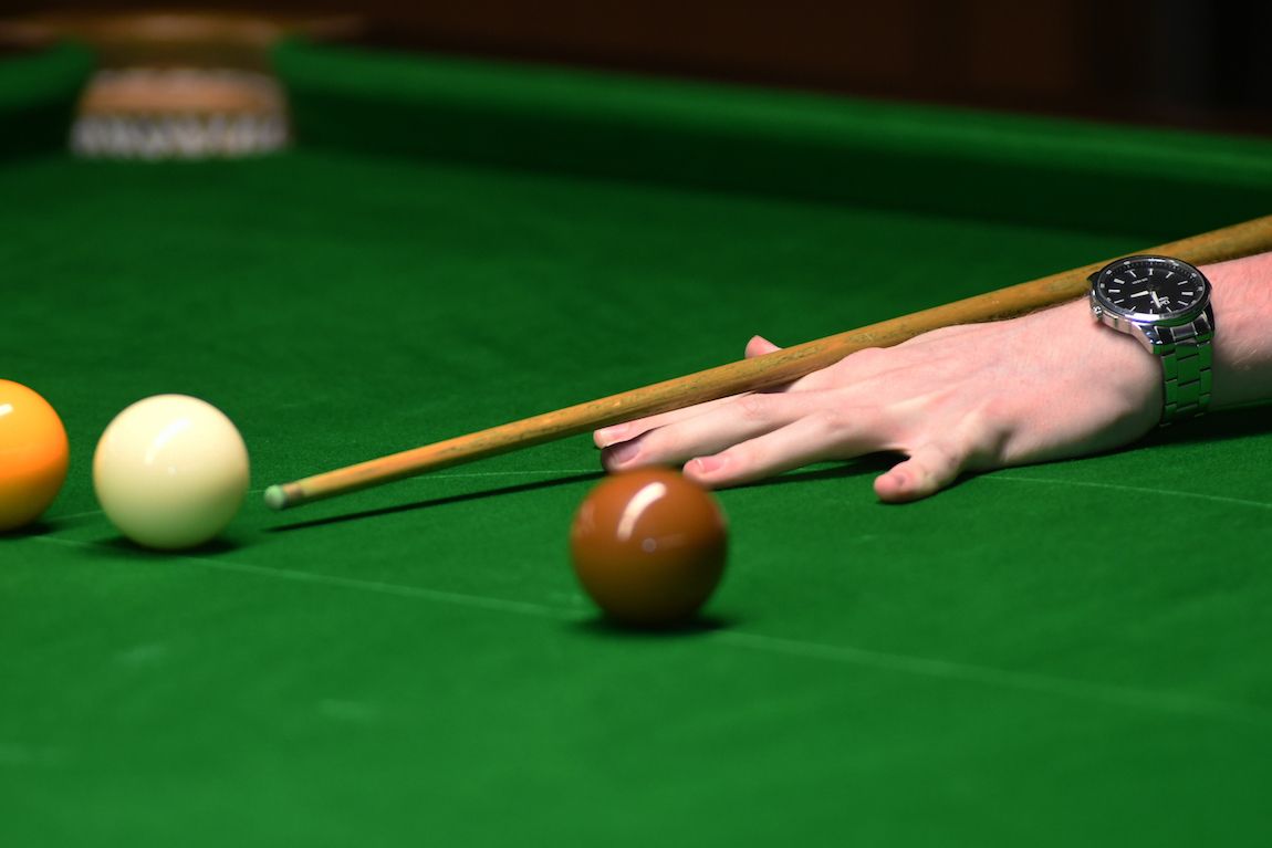 Een hand die snooker speelt, met de keu gericht op een bruine bal, op een groene tafel van vilt.