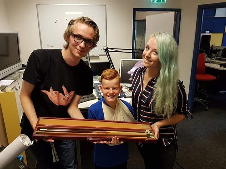 Three people, two adults and a child with an arm sling, hold a wooden case in an office setting.