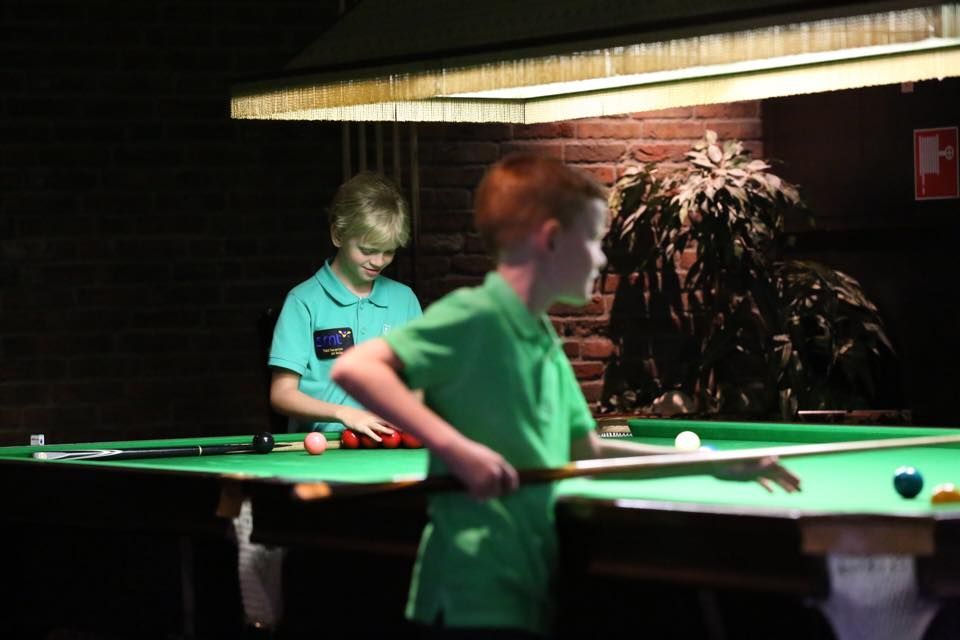 Twee jongens spelen pool in een schemerige kamer; de een mikt met een keu, de ander legt ballen op een plank.