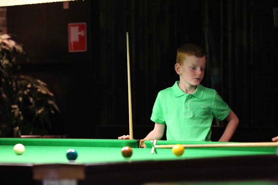 Jongen in groen shirt aan een pooltafel, met een keu in de hand. Ballen op het groene vilt, binnen.