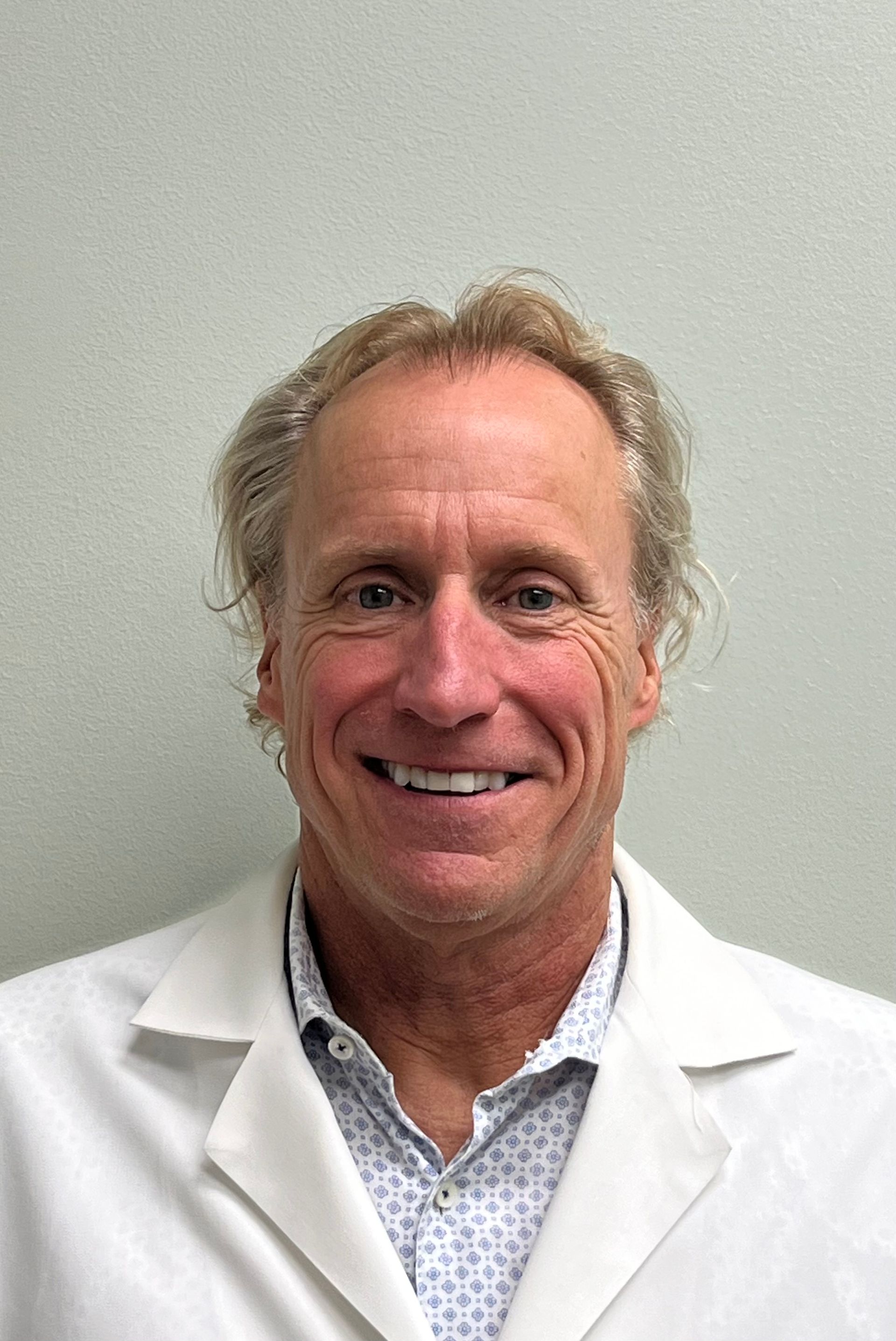 Man in a lab coat, smiling, wearing a blue shirt and patterned tie.