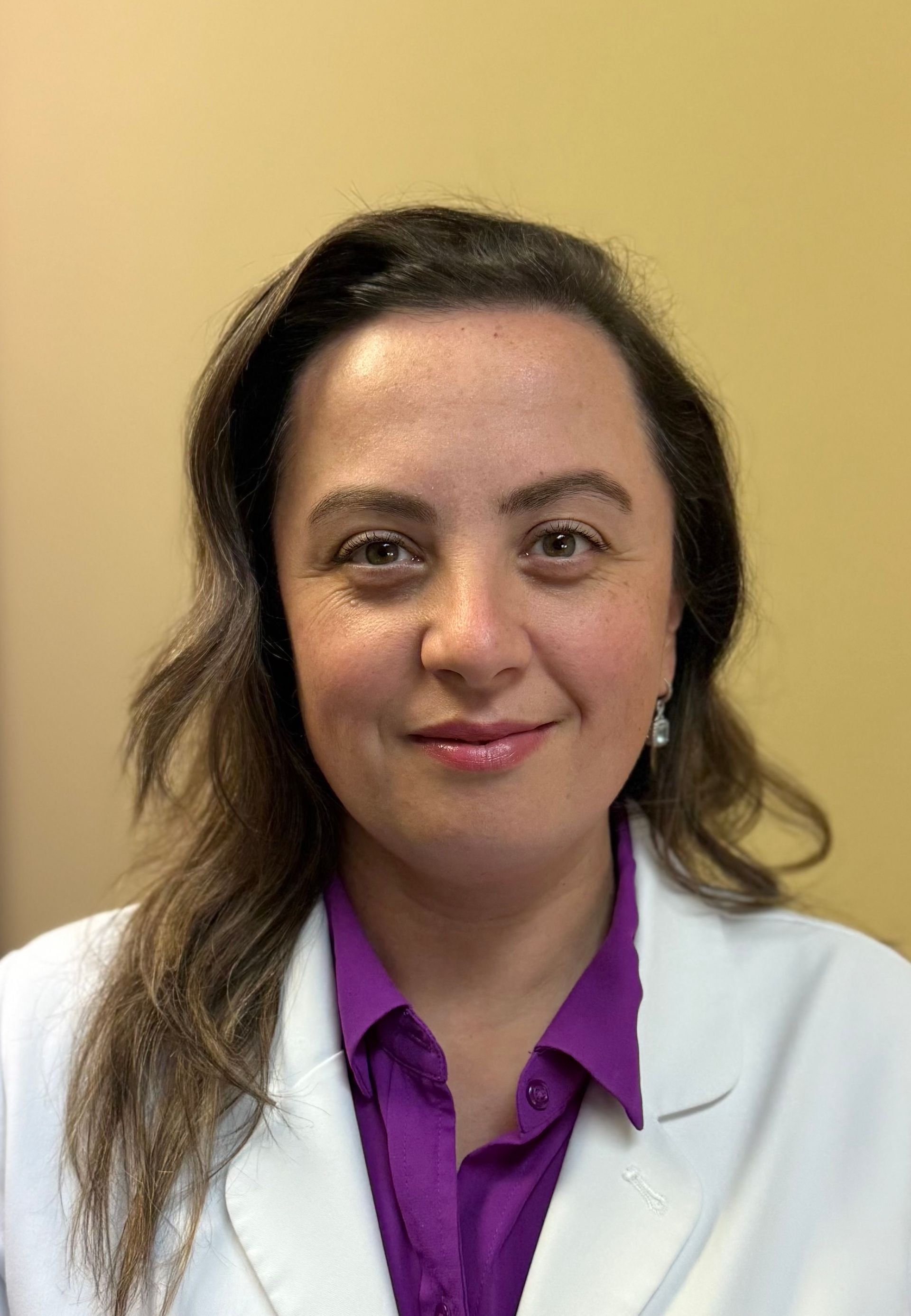 Woman in white coat, purple shirt, smiling, looking at the camera.