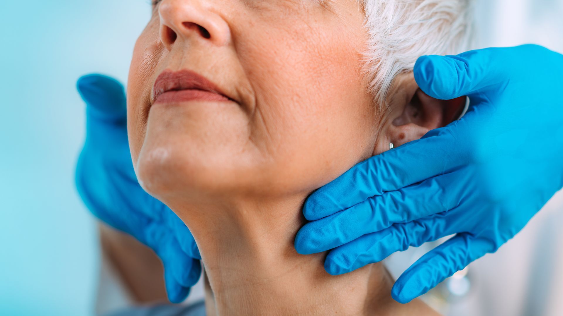 Medical professional in blue gloves examining an older person's neck. Close-up, light skin, healthcare setting.