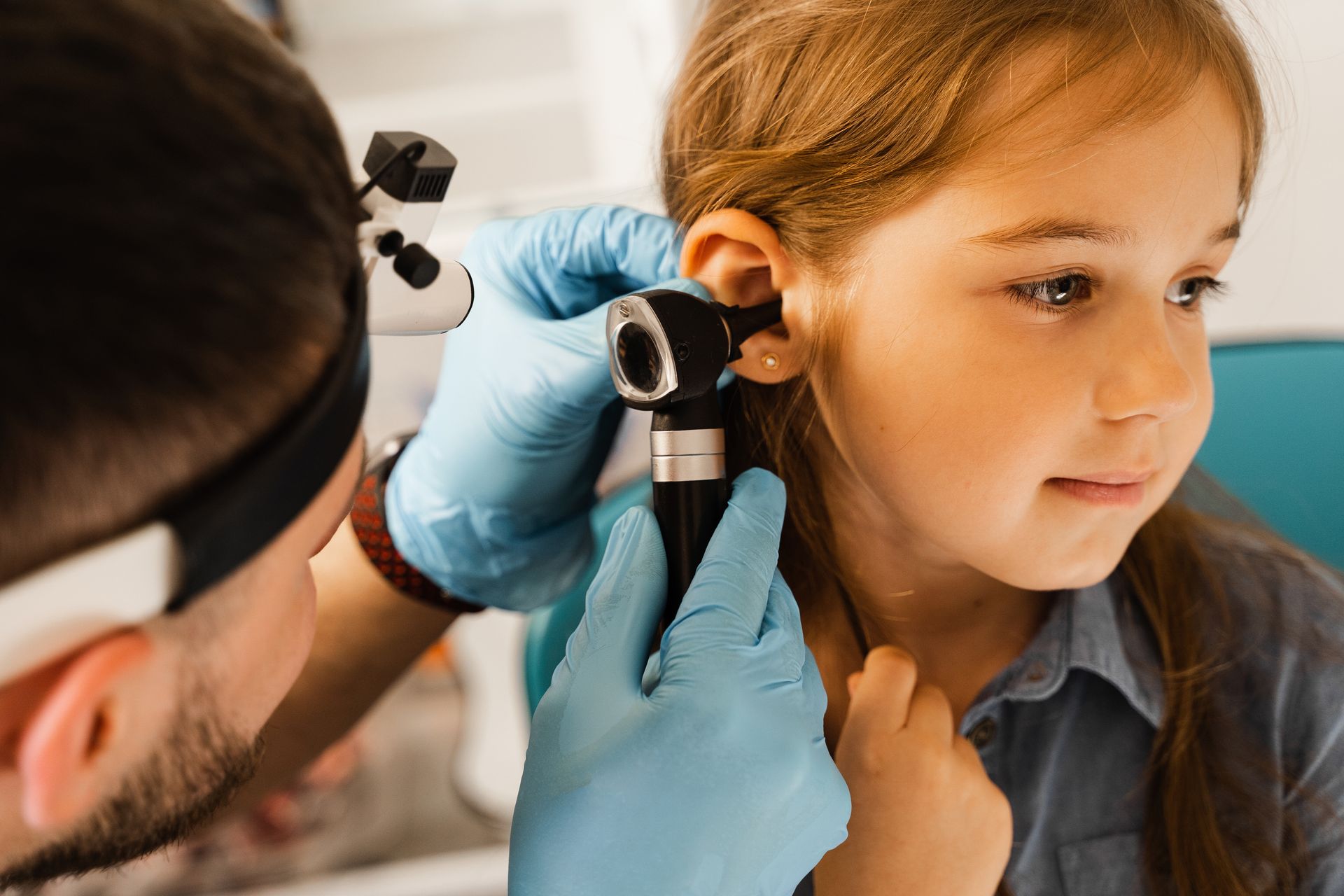 Doctor examining a child's ear with an otoscope. The child is seated, looking towards the doctor.
