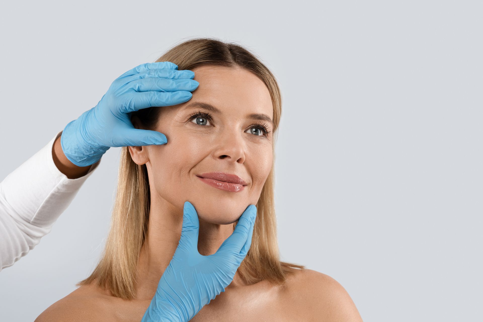 Woman's face being examined by gloved hands. Focus on forehead and cheeks, with a light blue background.