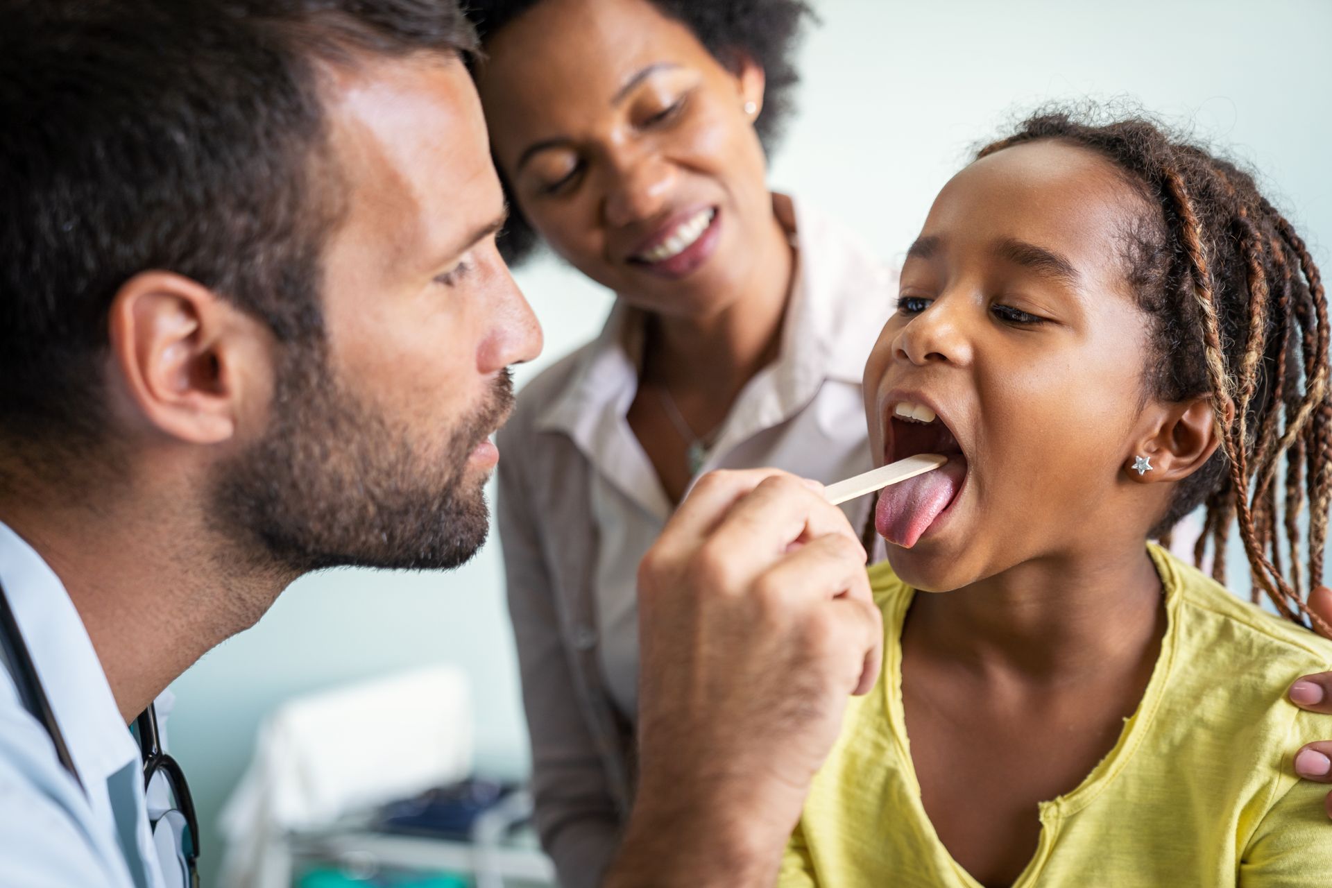 Doctor examining a child's throat with a tongue depressor, mother watching. Bright room, yellow shirt.