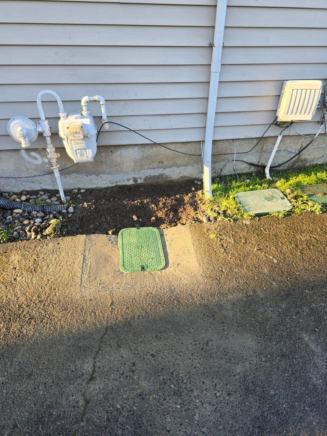 Side of house with gas meters, utility pipes, and a driveway with green utility covers.