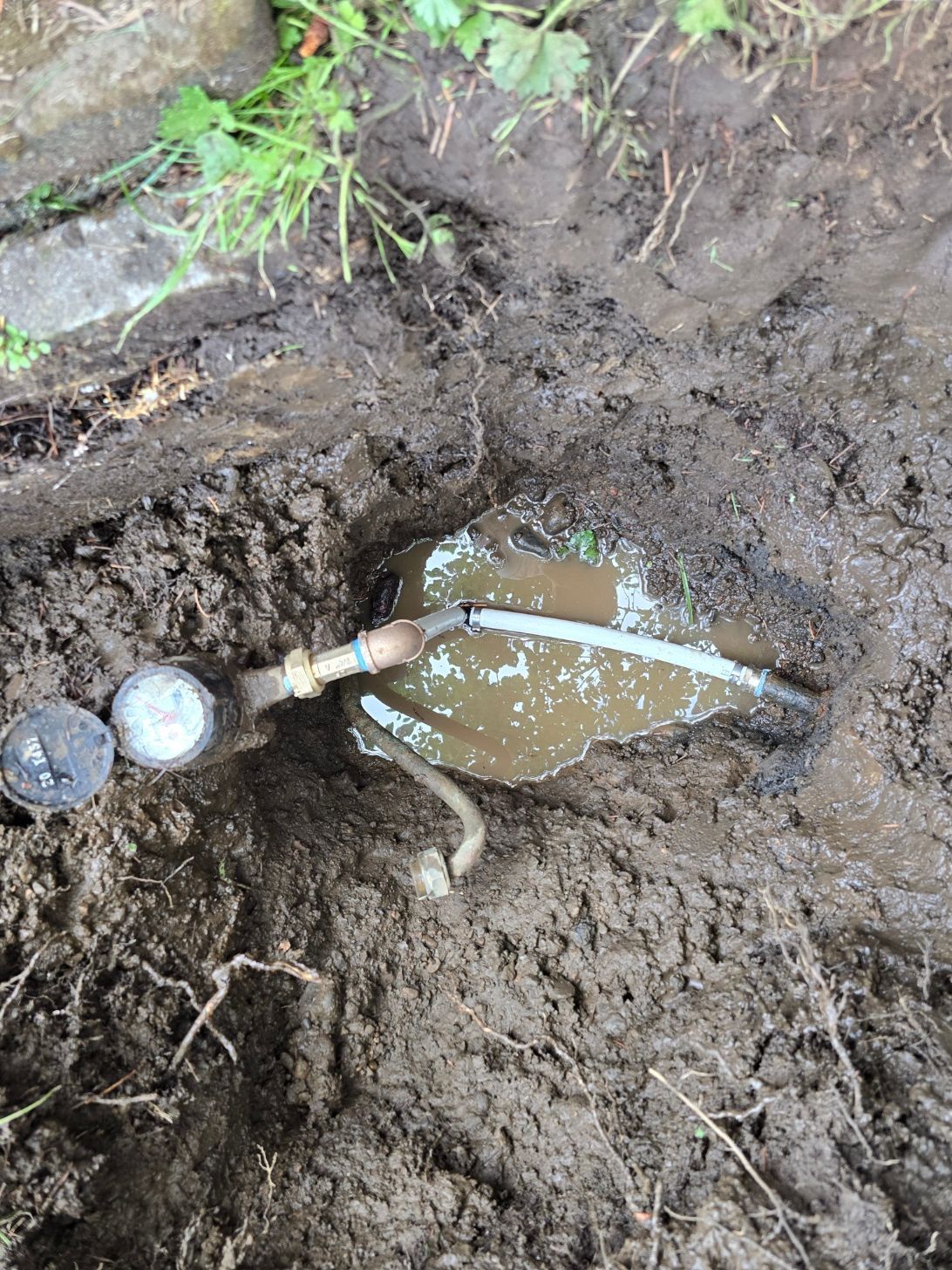 Muddy ground with an exposed water pipe and a small puddle of brown water near grass