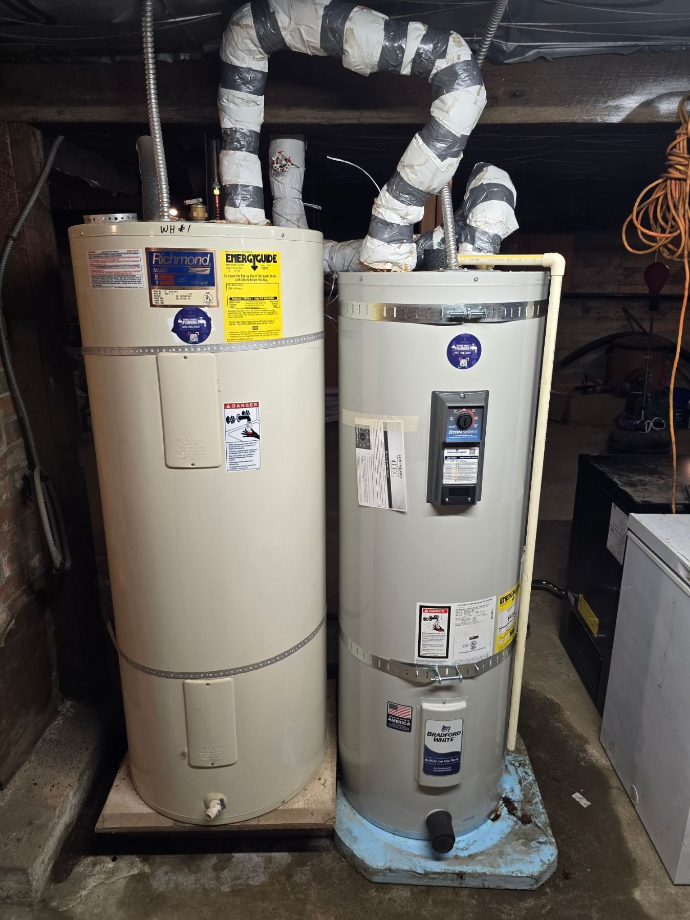 Two water heaters in a basement, one with foil-wrapped pipes and one on a blue drain pan.