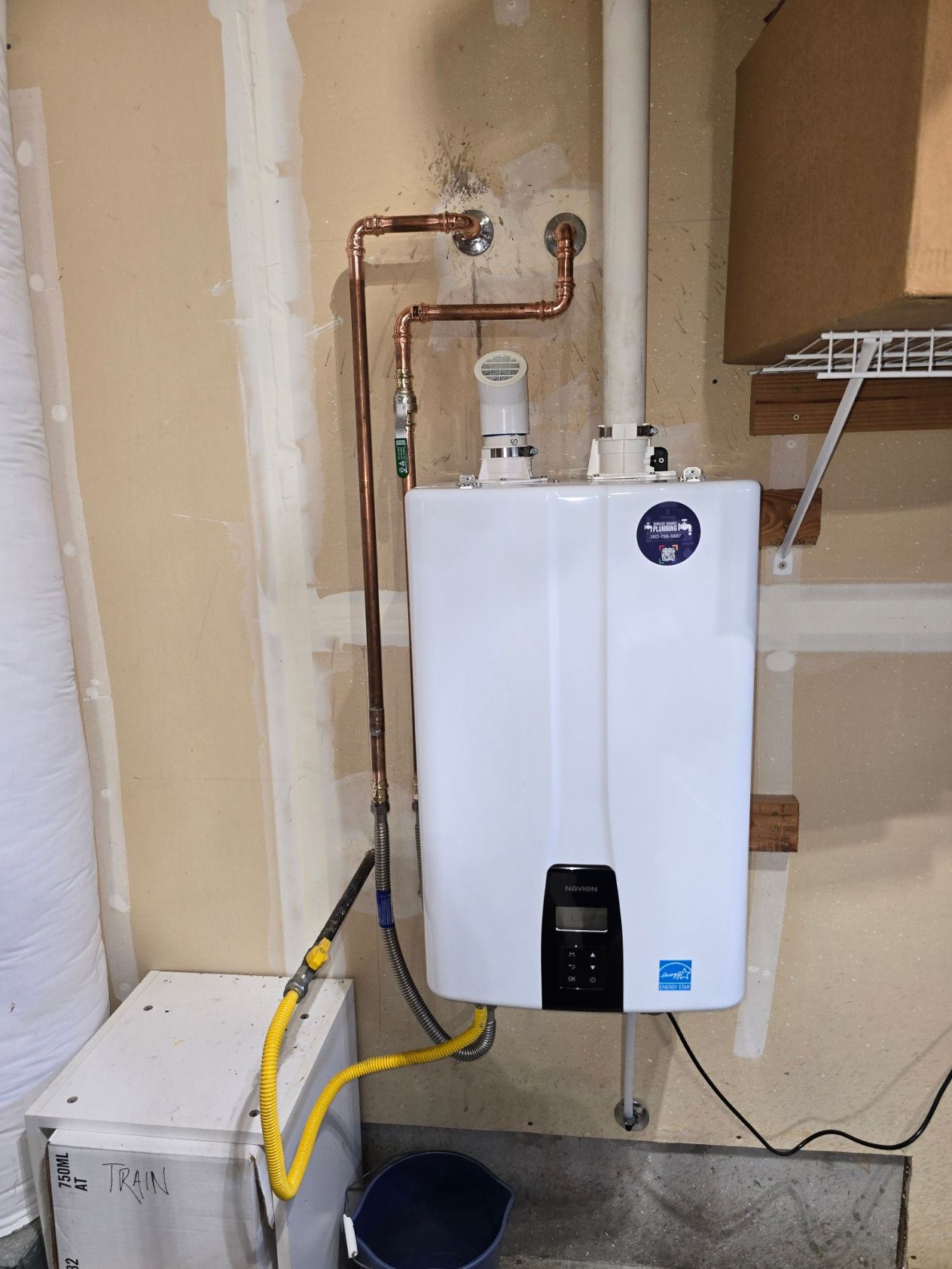 Water heater and wall-mounted boiler in a utility room with exposed copper pipes and a white storage bin.