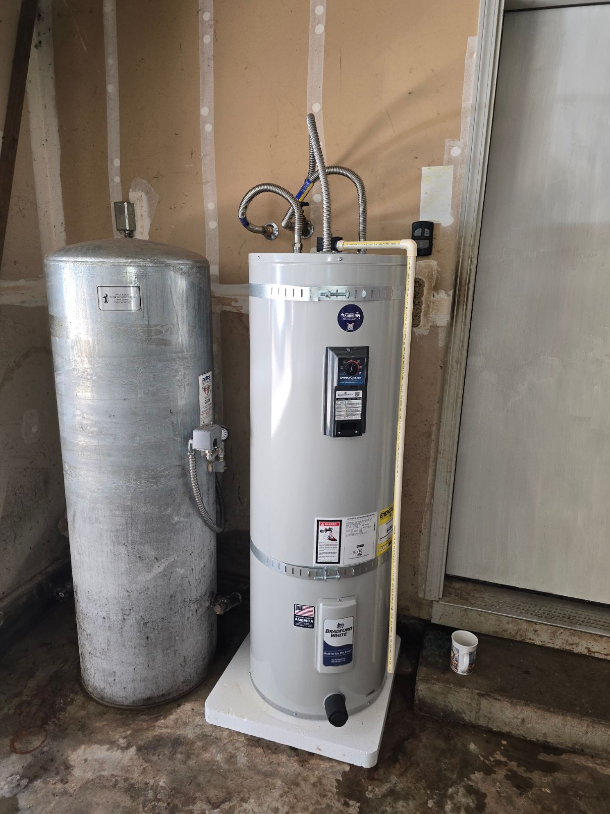 Utility room water heater next to a silver insulated tank, with exposed pipes and a door in the background